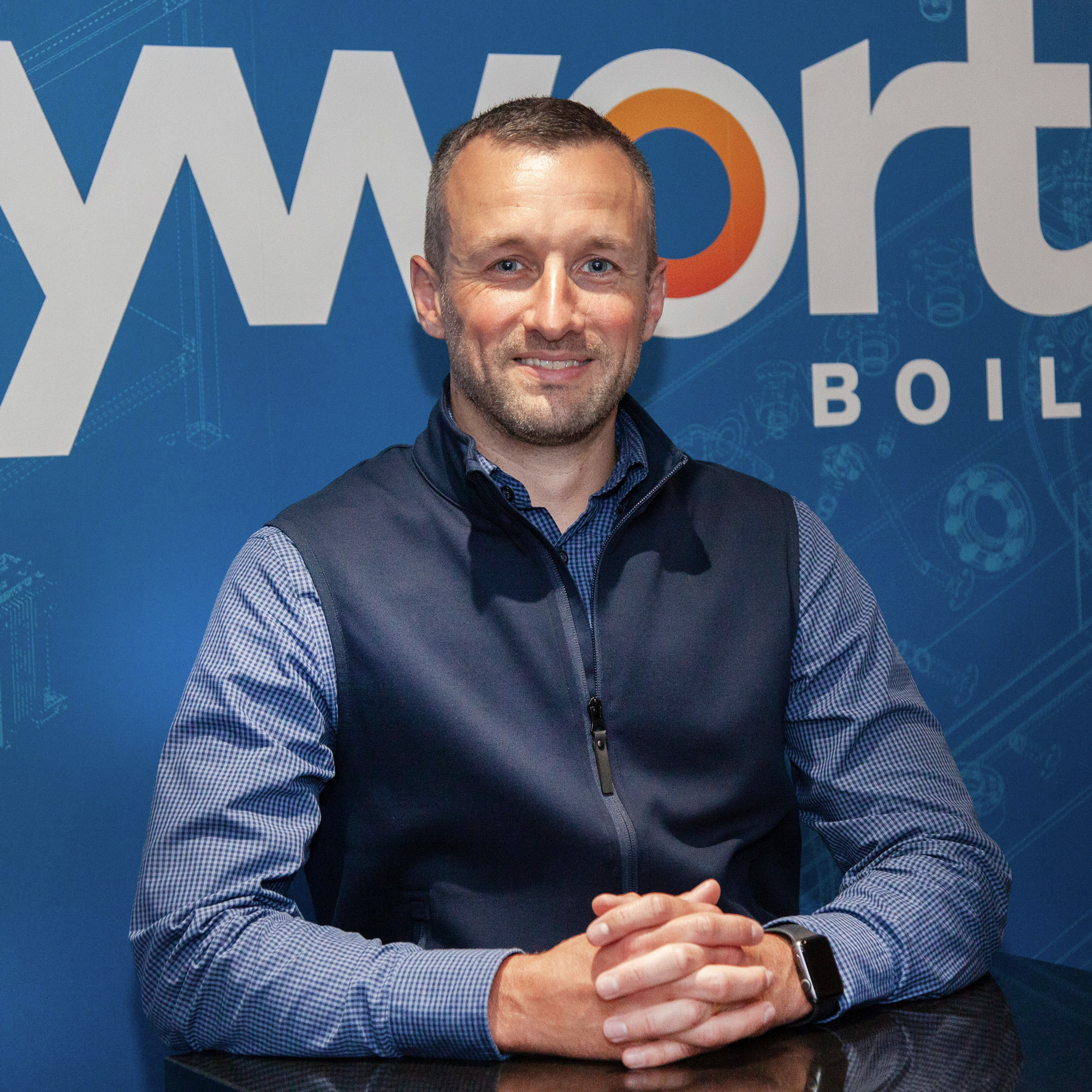 A man smiling at the camera with his hands folded in front of him. The background is blue 